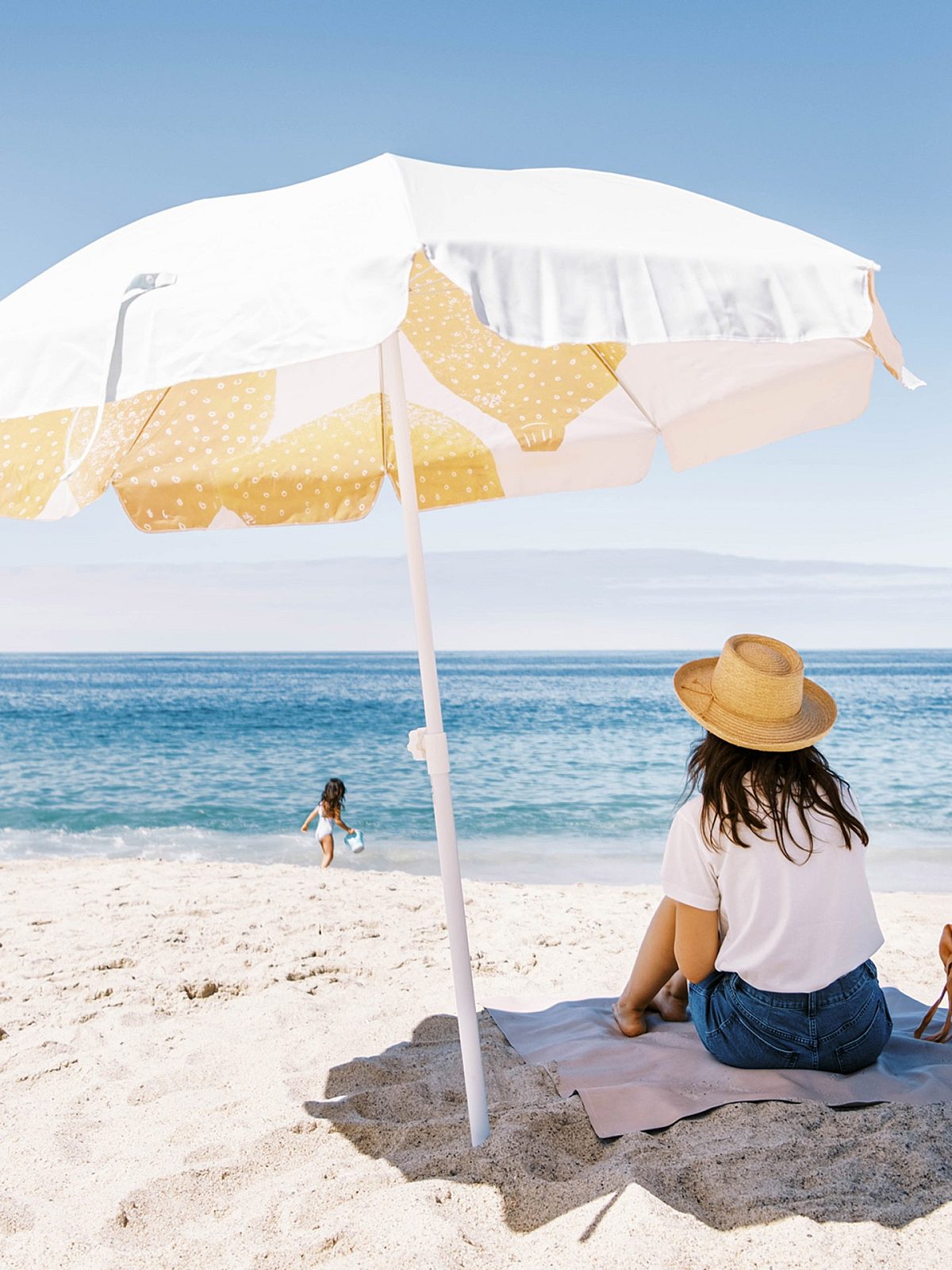 Woman watching a girl playing at the beach from her spot under a beach umbrella.